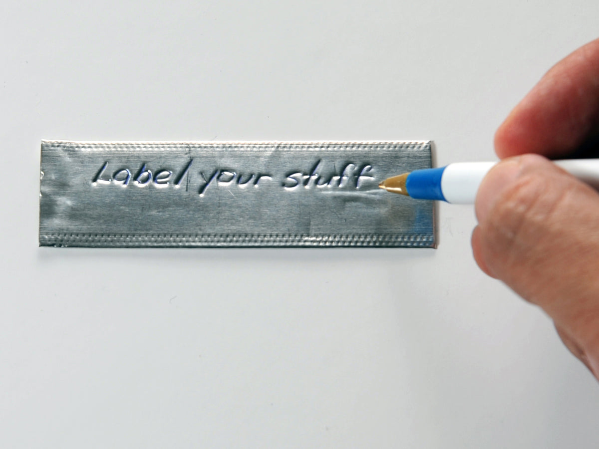 a hand holding a pen is shown above an emboss-o-tag that has been debossed with the words "Label your stuff" by the pen